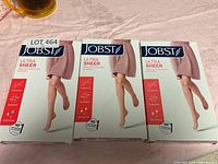 Three boxes of Jobst Ultra Sheer medical compression stockings shown side by side on a pink surface, showing front packaging with leg image and product benefits.