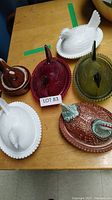 Five hen-shaped covered dishes displayed on wooden table: brown ceramic, red glass, green glass, white milk glass and brown/turquoise pheasant lusterware