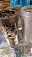 Photo showing vintage Coleman camp stove with metal top burner and fuel tank, placed next to a metal case and wrench on a table.