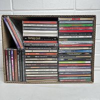 Photo of cardboard box filled with 50 assorted music CDs with visible titles and jewel cases.