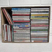 Box lot of 50 assorted compact discs stacked tightly in a cardboard box, showing several holiday-themed titles.
