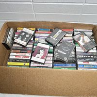 An overhead view of the cardboard box filled with a variety of vintage music cassette tapes mostly stacked in rows and some scattered on top.