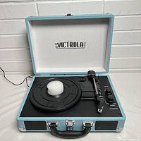 Front view of the portable record player opened, showing turntable, tonearm, control knobs, built-in speaker, and Victrola logo inside the lid.