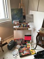 Photo showing wooden easel with textured painted canvas and a blank canvas board with a sketch, acrylic paint tubes, paint brush set, colored pencils, crayons, palettes, and an art supply starter kit on the floor nearby.
