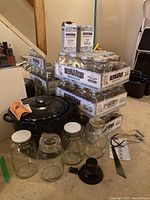 Photo showing three cartons of Bernardin 1L canning jars stacked, multiple loose large glass jars with lids, black granite ware pot with lid, funnel, jar lifter, and tongs on carpeted floor.