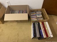 Two open cardboard boxes with numerous music CDs, one box with stacked jewel cases, the other with boxed set collections of various artists