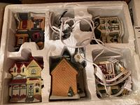 Top view of five ceramic Christmas village buildings in original styrofoam packaging with visible illumination cords and plugs.