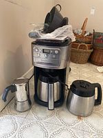 Photo showing Cuisinart DBG-900BCC drip coffee maker with built-in grinder and control panel, alongside a stainless steel thermal carafe, a glass coffee pot with black handle, and a small stainless steel stovetop espresso maker.