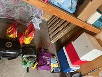 Image showing two Coleman coolers (red and blue), white styrofoam cooler on top, UniFlame portable black BBQ grill, three bags of charcoal, grill basket, charcoal lighter, purple bag, and folded tarp in a garage setting.