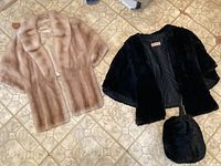 Overview of both fur stoles on tiled floor: light beige mink stole at left, black fur stole and matching muff at right