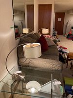 Pair of metal lamps on glass side table beside a patterned couch in basement. Lamps feature curved metal arms and perforated metal mesh cylindrical shades with plastic inner lining.