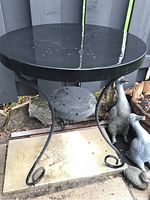 Full view of the black metal side table outdoors showing the round perforated top and curved legs with curled ends, set against a fence and garden decor.