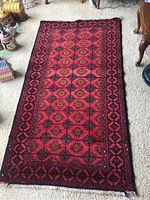 Full view of the rectangular Persian rug on a beige carpeted floor, showing the overall red and black geometric pattern and fringe.