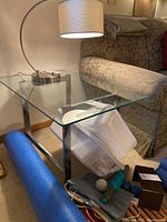 Full side view of one glass top side table with metal frame, showing the curved lamp and upholstered sofa in the background.