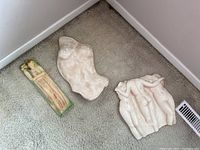 Three cast plaster wall art pieces pictured on carpet floor corner: painted rectangular floral woman piece, nude torso piece, and three nude figures piece.