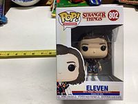 Funko Pop! Television Stranger Things #802 Eleven vinyl figure in original packaging with measuring tape beside it indicating size.