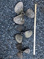 Three brass boat propellers arranged vertically on gravel next to a 36-inch yardstick for scale. Each propeller has three blades and shows signs of use and patina.