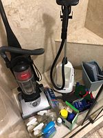 Wide shot of cleaning lot showing upright Hoover vacuum, Conair steam cleaner, multiple spray bottles, and containers of cleaning agents with accessories.
