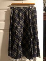 Mid-length vintage pleated skirt with navy, grey, beige plaid pattern hanging on wooden hanger in front of white door.