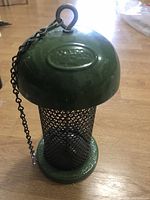 Full view of the hanging bird feeder, showcasing green ceramic top and base with metal mesh tube body and hanging chain.