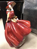 Side view of Royal Doulton porcelain figurine in red dress and red hat with blue ribbon holding a cream shawl.