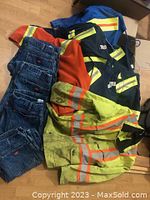 Work jeans and safety shirts with high-visibility reflective striping and different colors shown spread on floor.