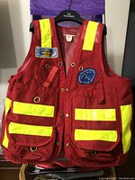 Front view of red surveyor vest showing multiple pockets, grommets, reflective yellow strips, and patches.