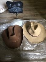 Top view of two cowboy hats, one brown felt and one beige felt, laid on a plastic sheet with a sign indicating lot number 69.
