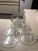Set of 4 clear Federal glass luncheon plates each with an embossed swirling leaf motif pattern and a large clear punch bowl with ladle, arranged on a kitchen counter.