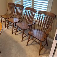 Four medium stained solid oak chairs lined up, with one larger captain chair at the front with armrests and three side chairs without armrests.