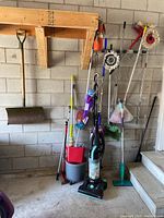Photo of cleaning and outdoor tools against cinder block wall including snow shovel, mops, buckets, vacuum cleaner, and steam mop