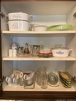 Cabinet shelves with multiple kitchenware items including Corning Ware casseroles, glassware, sauce servers, plates, and corn holders