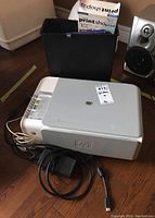HP Photosmart C3180 all-in-one printer with power and data cables, black bin behind, Print Shop software box on top of bin, on a wood floor.