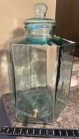 Full view of the transparent thick square glass jug with brassy valve near base and measuring tape showing approx 9 inches wide base.