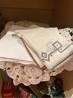 Photo of two white fabric napkins, one with geometric embroidered pattern in brown and blue tones, placed on a crocheted pink lace item