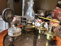 Photo showing three oil lamps on glass table including the clear glass base lamp with chimney and two brass base oil lamps, one with hinged lid open revealing wick