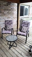 Two steel patio chairs with purple-grey patterned cushions and lumbar pillows on wooden deck, near house wall.