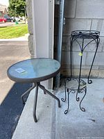 Overall set of glass-topped metal patio table and two wrought iron plant stands shown outside near garage.
