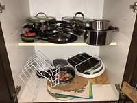 Kitchen cabinet shelf with multiple assorted pots, pans with lids, baking trays, cutting boards, and food chopper parts arranged on two shelves.