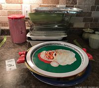 Stack of clear and colored glass bakeware dishes, Christmas trays with Santa design, and a red striped metal tin on counter