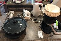 Set of small kitchen appliances on countertop including a Chefman electric skillet with glass lid, white electric kettle, two white Black & Decker hand mixers with beaters, and a black Sunbeam coffee maker loaded with filters.