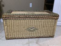 Photo showing the entire large wicker chest or blanket box on floor with beige wall background.