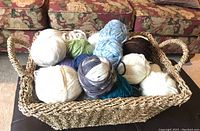 Woven rope basket filled with assorted yarn balls in various colors and textures, shown from above on dark surface.