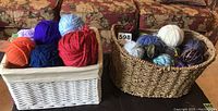 Two baskets side by side on a table, the white rope basket on the left filled with colorful yarn balls including orange, navy blue, red, light blue, purple, and lavender. The natural wicker basket on the right filled with various yarn colors including blue, white, multi-tone blues and browns, and gray.
