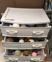 White plastic storage drawers unit with three drawers partially open showing multiple colored yarn balls inside.