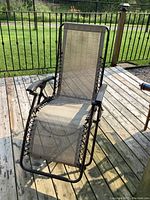 Zero gravity lounger chair on wooden deck, beige mesh fabric seating, black metal frame, showing front view and armrests.