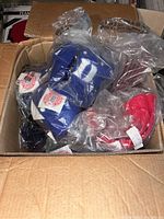 Box with multiple sealed NCAA team color ball caps in blue, black, red, and maroon shades.