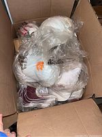Box of multiple white adjustable baseball caps individually wrapped in plastic. Various NCAA team logos visible including Clemson and Indiana University.