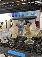 Image of assorted art glass on metal shelving including vases in cream, blue, multicolor, a whale figurine, a glass cake dish, and a lidded vessel, showcasing varied shapes and textures.
