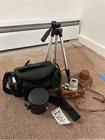 Lot items on carpet floor showing Argus camera with leather case, black camera bag, leather lens case, and Vivitar tripod.